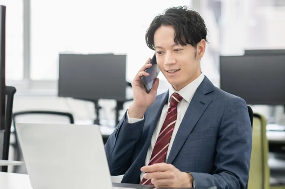 スーツを着た男性がPCを見ながらスマホで話をしている様子