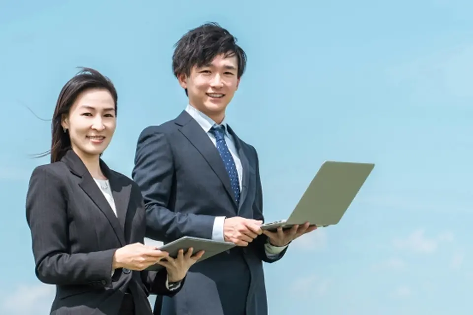 PCを持って立っているスーツを着た男性と女性が笑顔でこちらを見ている様子